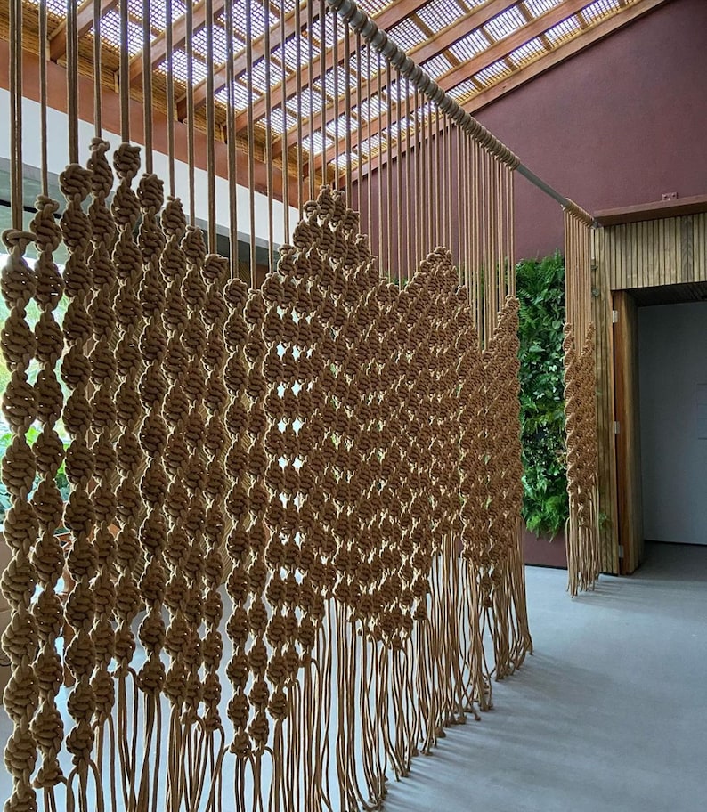 May include: A large macrame wall hanging made of natural brown rope. The wall hanging is made of a series of knots and is designed to resemble a curtain. The wall hanging is hanging in front of a wooden wall.