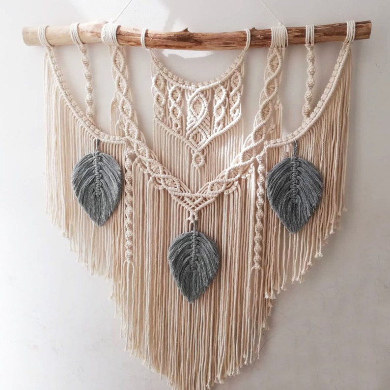 May include: A macrame wall hanging with a natural wood dowel and three gray feather accents. The wall hanging is made with white cotton cord and has a bohemian style.