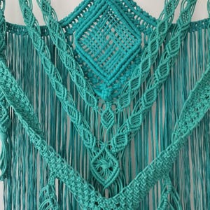 Op de afbeelding: Een turquoise macram&eacute; wandhanger met een ruitpatroon in het midden en franjes langs de onderkant.