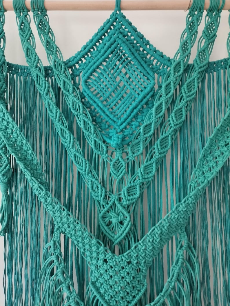 Op de afbeelding: Een turquoise macram&eacute; wandhanger met een ruitpatroon in het midden en franjes langs de onderkant.