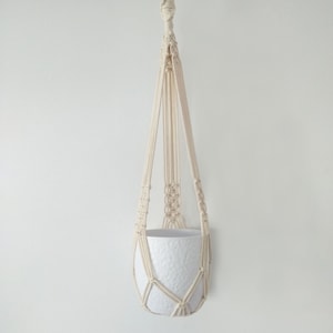 May include: A white macrame plant hanger with a white ceramic pot. The hanger is made of cotton cord and has a simple design.