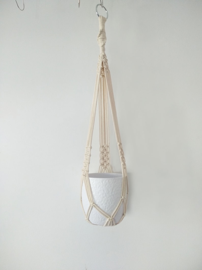May include: A white macrame plant hanger with a white ceramic pot. The hanger is made of cotton cord and has a simple design.