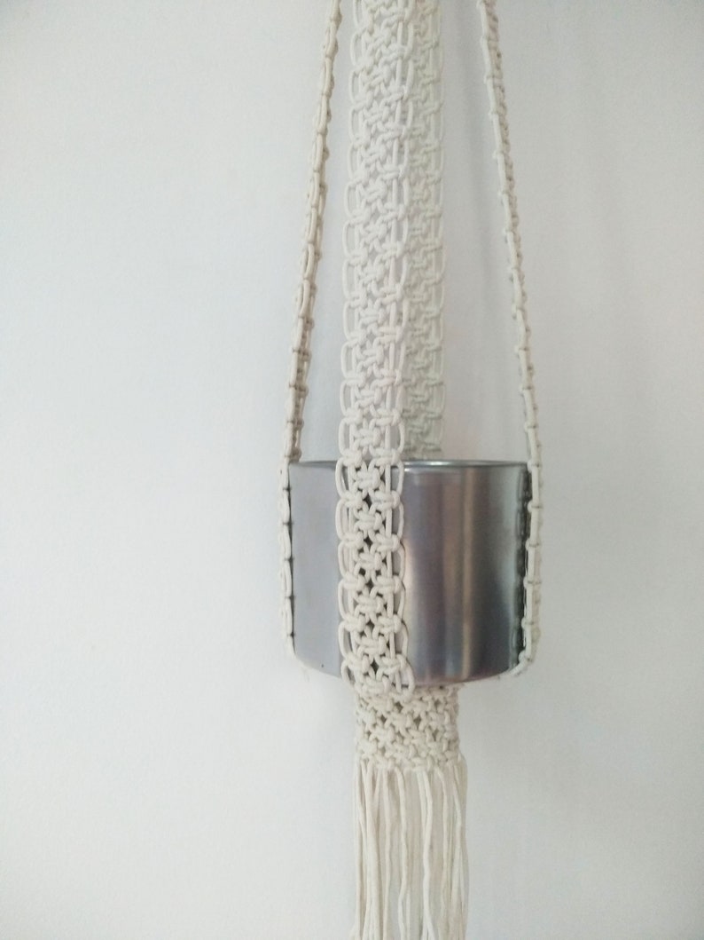 Pu&ograve; includere: Un portavasi in macram&egrave; bianco con un vaso in metallo argentato. Il portavasi ha una frangia sul fondo.
