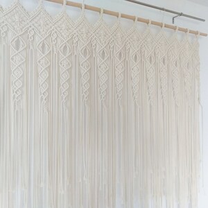May include: Cream-colored macrame wall hanging. The top features a geometric design, with vertical strands cascading down. The piece is suspended from a wooden dowel. The wall hanging is a decorative textile element.