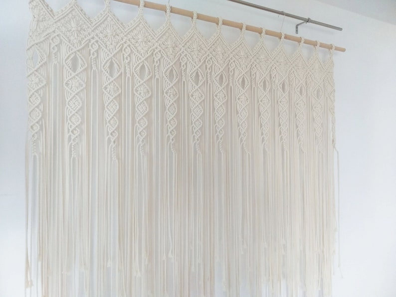 May include: Cream-colored macrame wall hanging. The top features a geometric design, with vertical strands cascading down. The piece is suspended from a wooden dowel. The wall hanging is a decorative textile element.