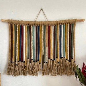 Op de afbeelding: Een wandhanger gemaakt van verschillende gekleurde garenstrengen met zwarte uiteinden en een jute franje. De garenstrengen zijn met elkaar verweven en hangen aan een houten lat.