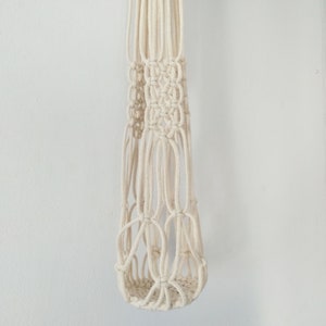 May include: A white macrame plant hanger with a knotted design. The hanger is made of cotton cord and has a cylindrical shape.