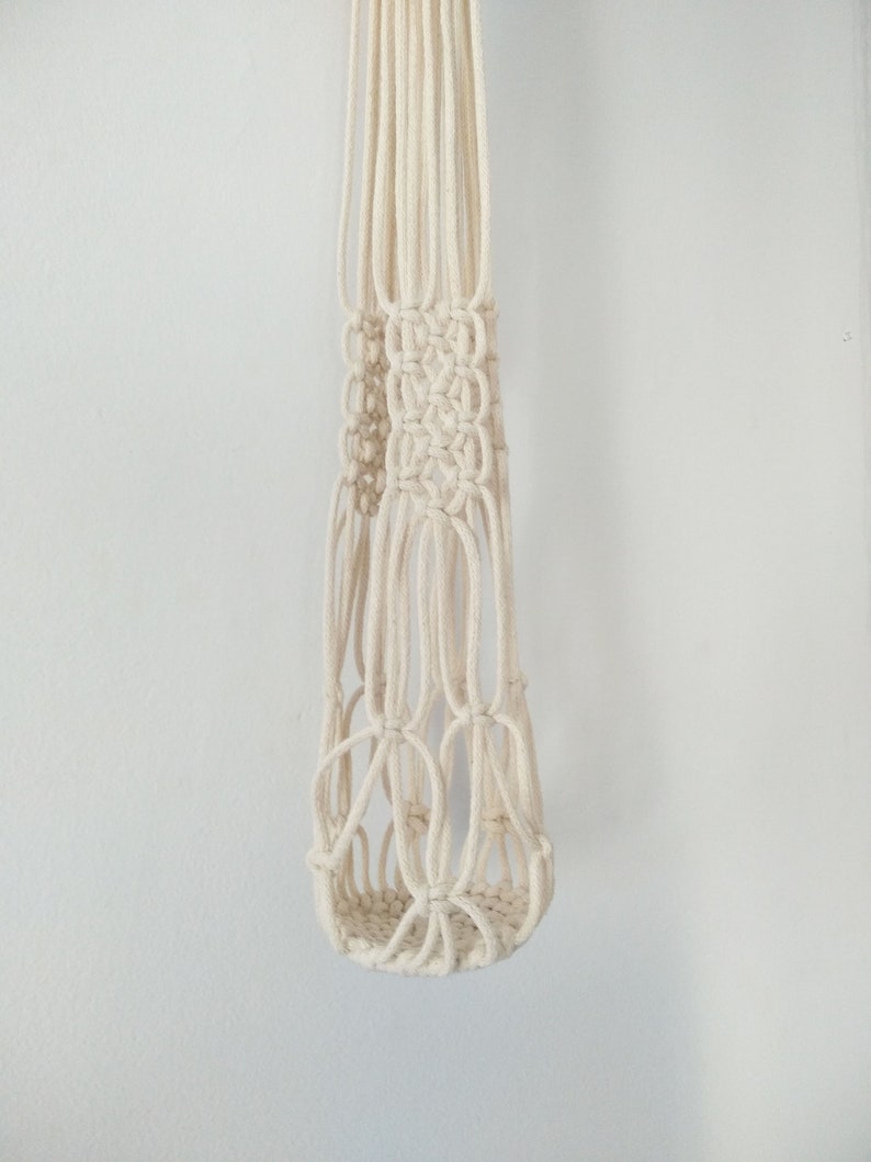 May include: A white macrame plant hanger with a knotted design. The hanger is made of cotton cord and has a cylindrical shape.