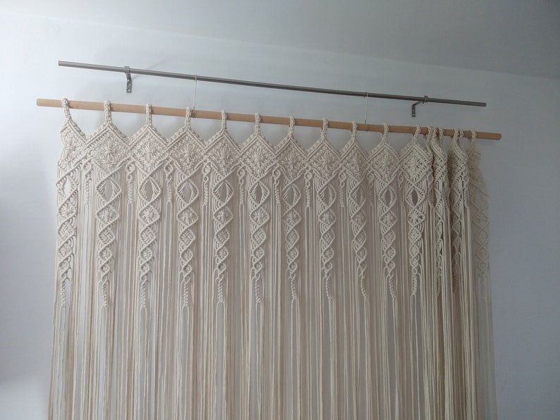 May include: Cream-colored macrame curtain hanging from a wooden rod and metal hardware. The curtain features a geometric design at the top with diamond and spiral patterns, and long, flowing fringe.