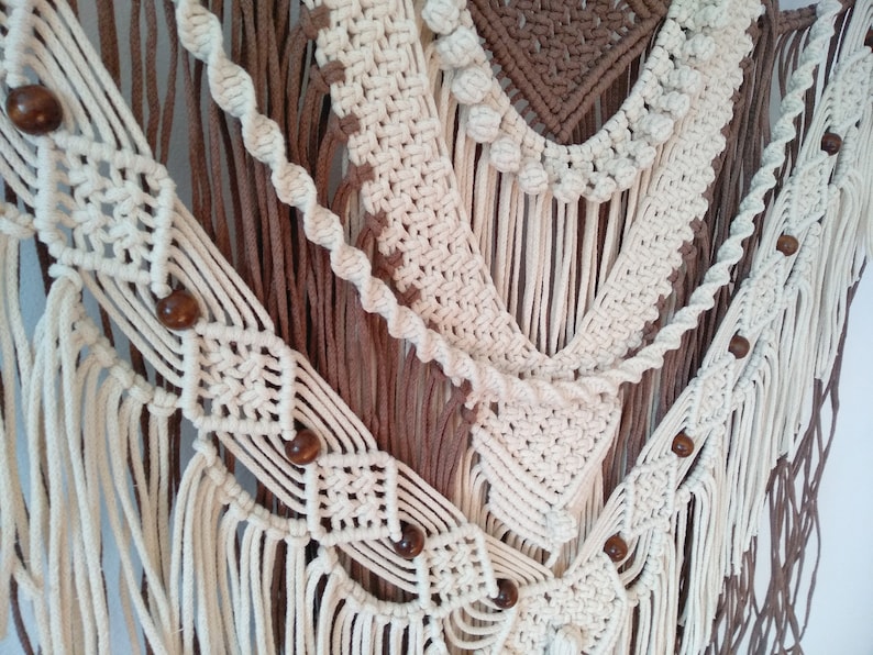 May include: A white and brown macrame wall hanging with wooden beads. The macrame is made with a variety of knots and patterns, and the beads are spaced evenly throughout the hanging.