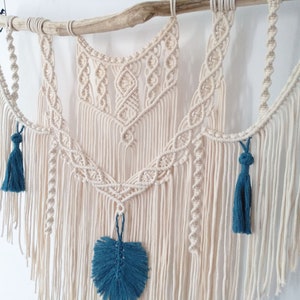 May include: A macrame wall hanging with a natural wood branch at the top. The macrame is made with cream-colored cord and features a geometric design. The hanging has three blue tassels and a blue leaf-shaped embellishment.