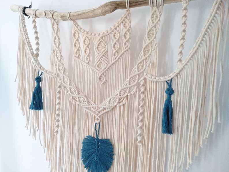 May include: A macrame wall hanging with a natural wood branch at the top. The macrame is made with cream-colored cord and features a geometric design. The hanging has three blue tassels and a blue leaf-shaped embellishment.