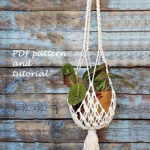 以下が含まれることがあります： 木製の壁に白いマクラメのプラントハンガーがタッセル付きで吊り下げられています。ハンガーには緑の葉の入った鉢植えの植物が入っています。画像には「PDF pattern and tutorial」というテキストが表示されています。