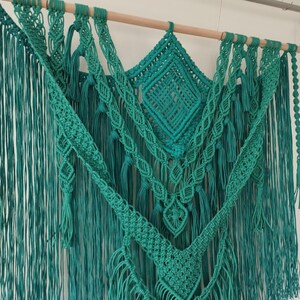Op de afbeelding: Een teal macram&eacute; wandhanger met verschillende knopen en texturen. De hanger is gemaakt van katoenen koord en heeft een houten lat aan de bovenkant.