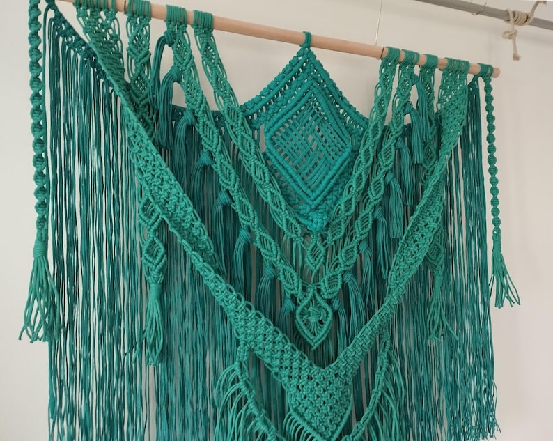 Op de afbeelding: Een teal macram&eacute; wandhanger met verschillende knopen en texturen. De hanger is gemaakt van katoenen koord en heeft een houten lat aan de bovenkant.