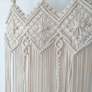 May include: A white macrame wall hanging with a geometric pattern. The hanging is made of cotton cord and has a wooden dowel at the top.