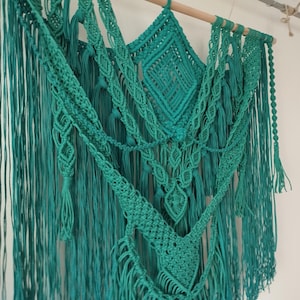Op de afbeelding: Een turquoise macram&eacute; wandhanger met verschillende knopen en texturen. De hanger heeft lange franjes en is gemaakt van katoenen koord.