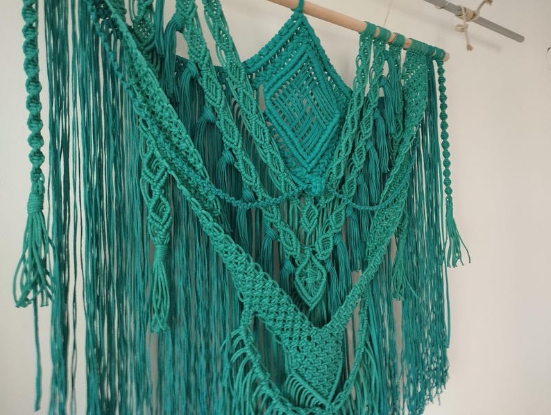 Op de afbeelding: Een turquoise macram&eacute; wandhanger met verschillende knopen en texturen. De hanger heeft lange franjes en is gemaakt van katoenen koord.