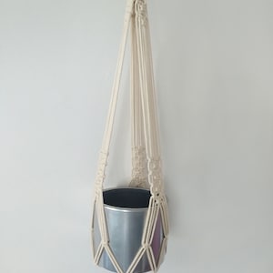 May include: A white macrame plant hanger with a silver metal pot hanging from it.