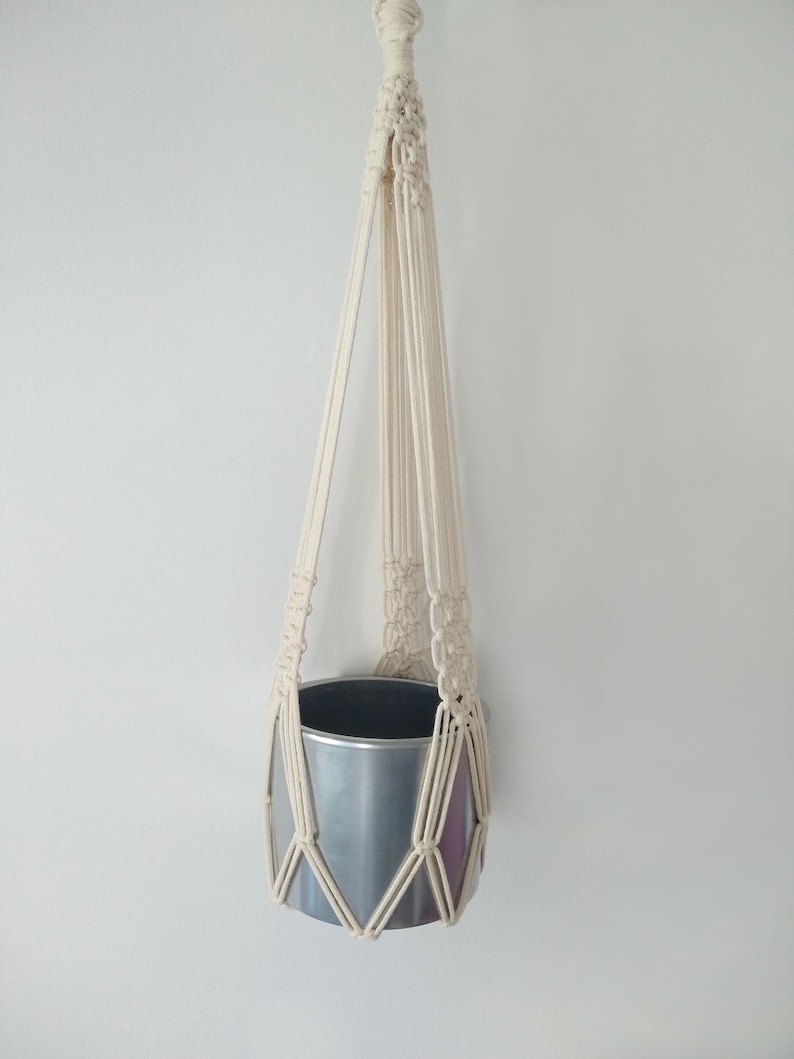 May include: A white macrame plant hanger with a silver metal pot hanging from it.
