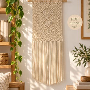 May include: A cream-coloured macrame wall hanging suspended from a wooden dowel, featuring a textured knot pattern with three central diamond-shaped geometric designs and long, straight fringe at the bottom. The image includes the text PDF tutorial.