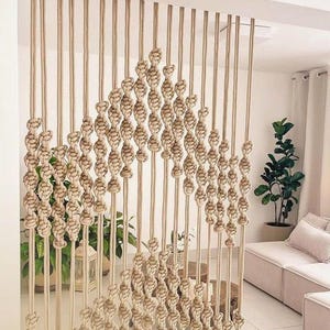 May include: A macrame wall hanging made of natural beige rope, with a diamond pattern and a fringe at the bottom. The hanging is in front of a white wall and a couch with beige cushions.