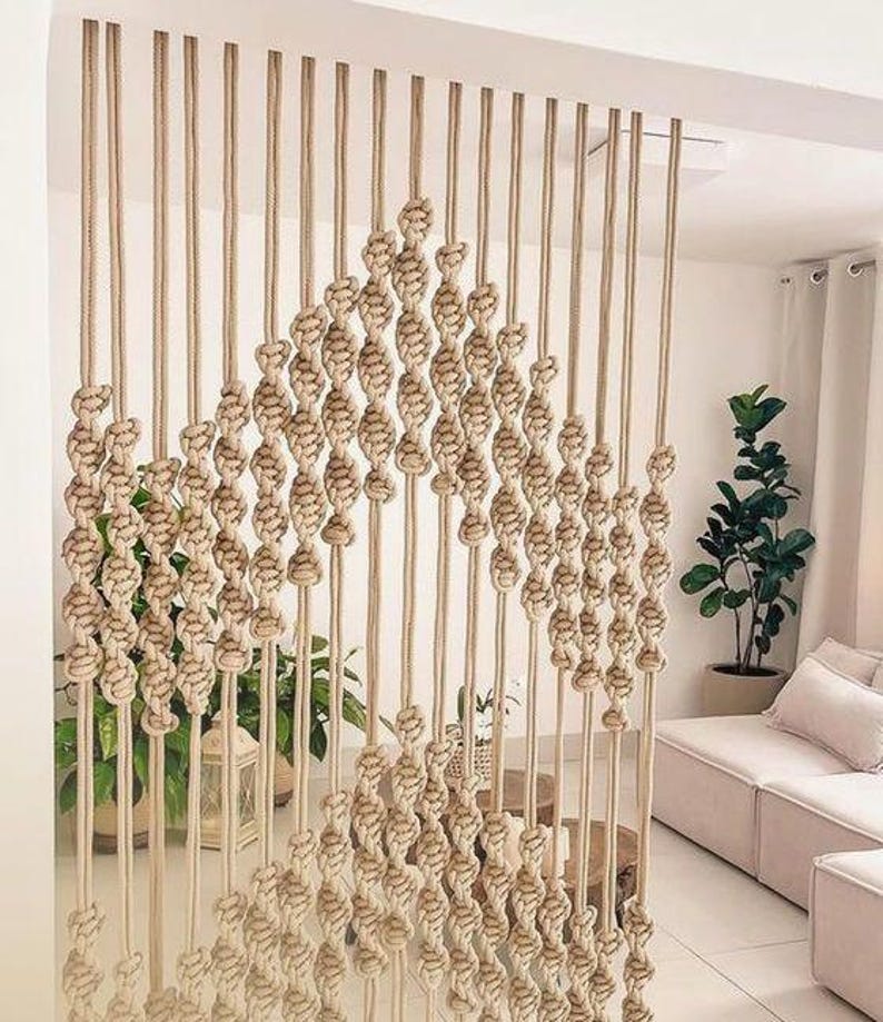 May include: A macrame wall hanging made of natural beige rope, with a diamond pattern and a fringe at the bottom. The hanging is in front of a white wall and a couch with beige cushions.