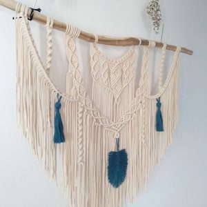 May include: A macrame wall hanging with a natural wood branch, featuring a geometric design with white cotton cord and teal blue tassels and a feather.