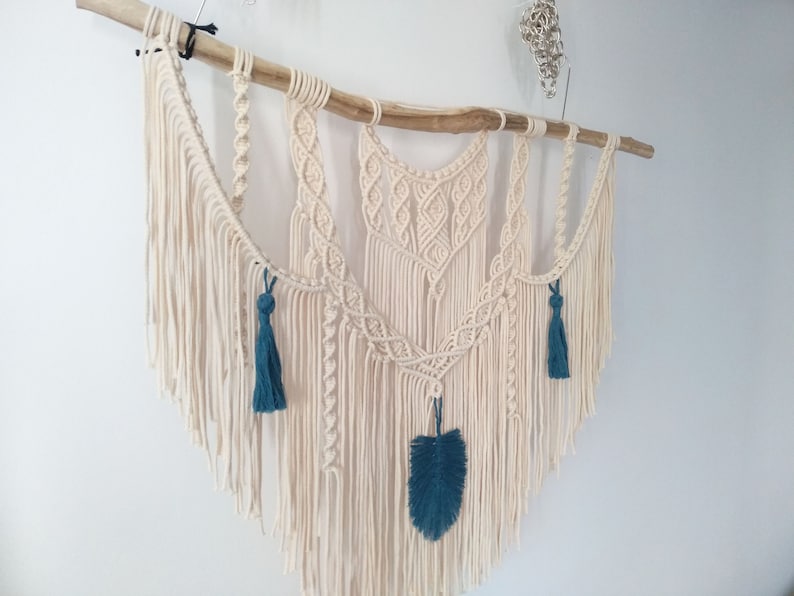 May include: A macrame wall hanging with a natural wood branch, featuring a geometric design with white cotton cord and teal blue tassels and a feather.