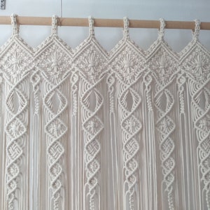 May include: Cream-colored macrame curtain with intricate diamond and spiral patterns. The curtain hangs from a wooden rod, showcasing a bohemian style. The design features knotted details and long, flowing fringe.