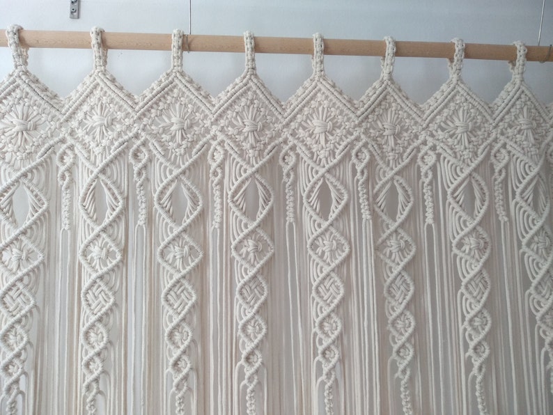 May include: Cream-colored macrame curtain with intricate diamond and spiral patterns. The curtain hangs from a wooden rod, showcasing a bohemian style. The design features knotted details and long, flowing fringe.