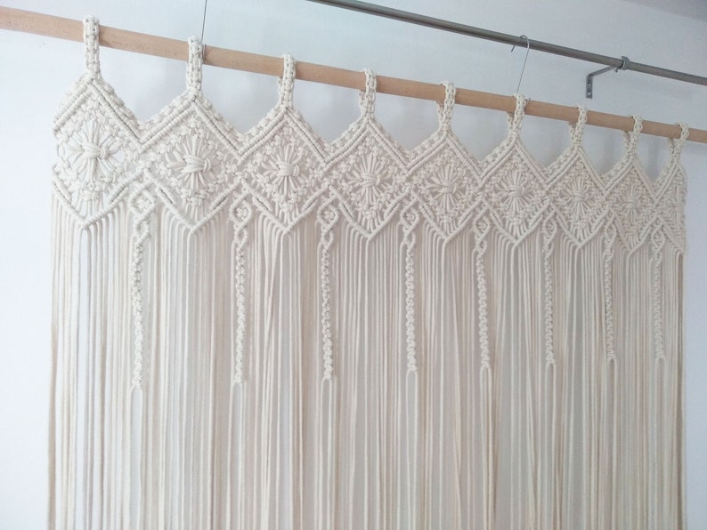 May include: A white macrame wall hanging with a geometric pattern. The hanging is made of cotton cord and is attached to a wooden dowel rod.