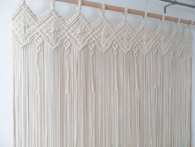 May include: A white macrame wall hanging with a geometric pattern. The hanging is made of cotton cord and is attached to a wooden dowel rod.