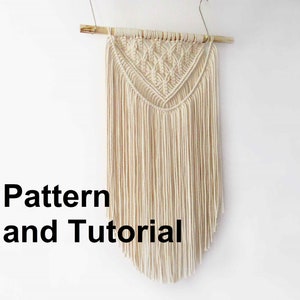 May include: A white macrame wall hanging with a geometric pattern and fringe. The hanging is attached to a wooden dowel rod.