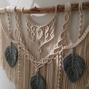 May include: A macrame wall hanging with a natural wood dowel and gray feather accents. The wall hanging features a geometric pattern with a bohemian style.