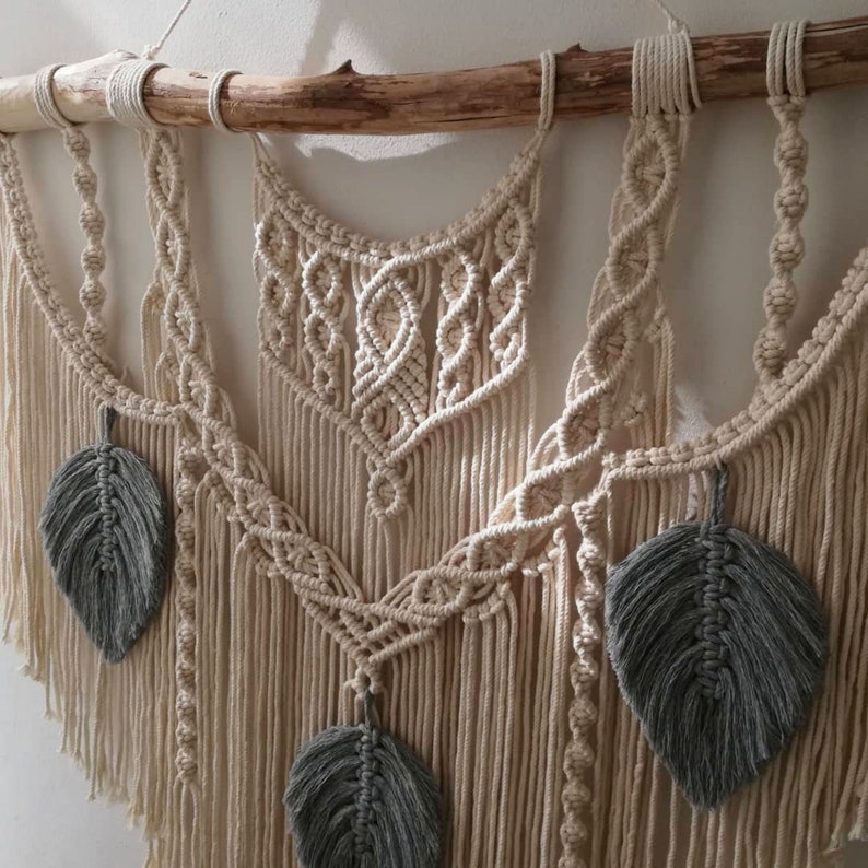 May include: A macrame wall hanging with a natural wood dowel and gray feather accents. The wall hanging features a geometric pattern with a bohemian style.