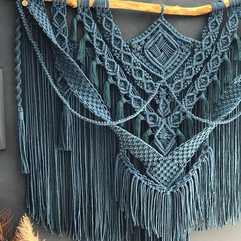 Op de afbeelding: Een tealblauwe macram&eacute; wandhanger met een geometrisch patroon en franjes. De hanger is gemaakt van katoenen koord en is bevestigd aan een houten lat.