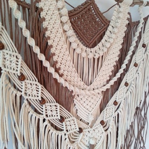 May include: A macrame wall hanging with a brown and white color scheme. The hanging features a variety of knots and textures, including fringe and beads.