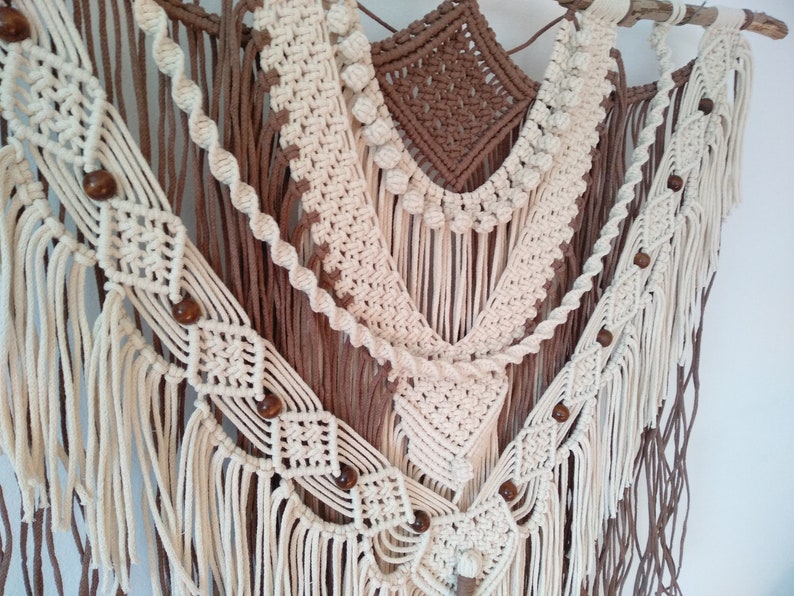 May include: A macrame wall hanging with a brown and white color scheme. The hanging features a variety of knots and textures, including fringe and beads.