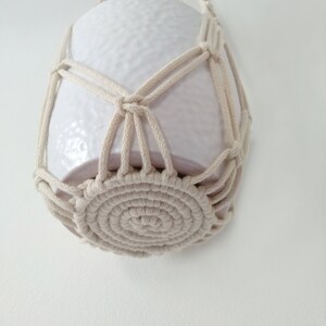 May include: A white ceramic planter hanging in a macrame plant hanger. The hanger is made of natural cotton rope and has a circular base.
