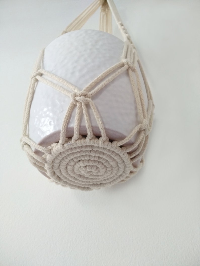 May include: A white ceramic planter hanging in a macrame plant hanger. The hanger is made of natural cotton rope and has a circular base.