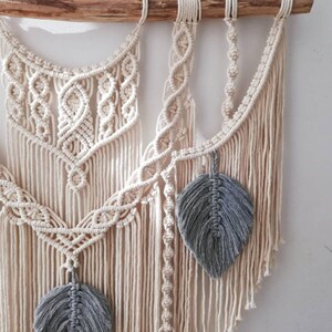 May include: A macrame wall hanging with a white cotton cord and gray feather accents. The wall hanging is made with a variety of knots and patterns.
