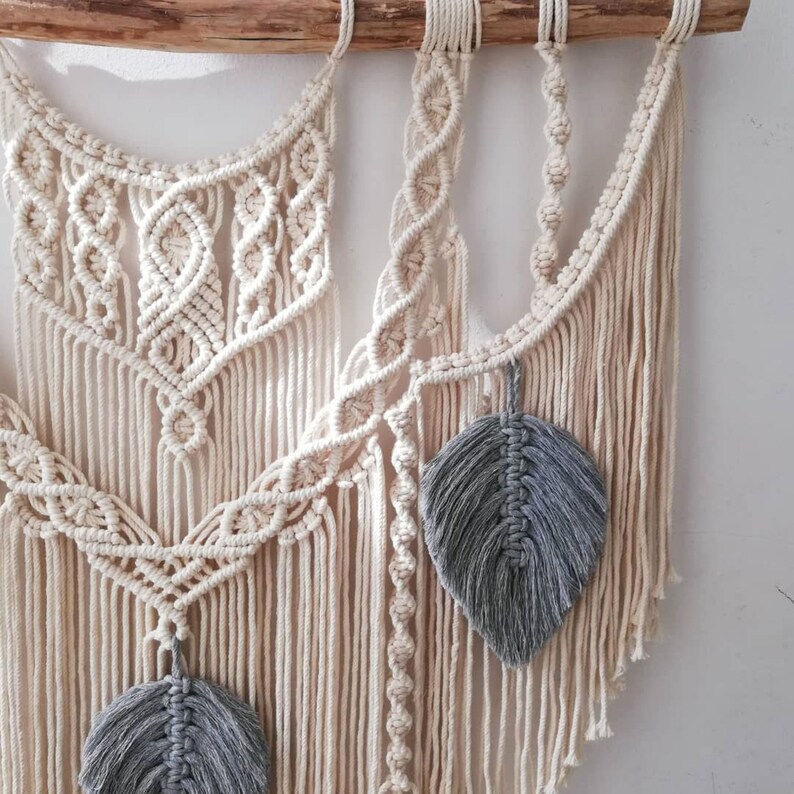 May include: A macrame wall hanging with a white cotton cord and gray feather accents. The wall hanging is made with a variety of knots and patterns.