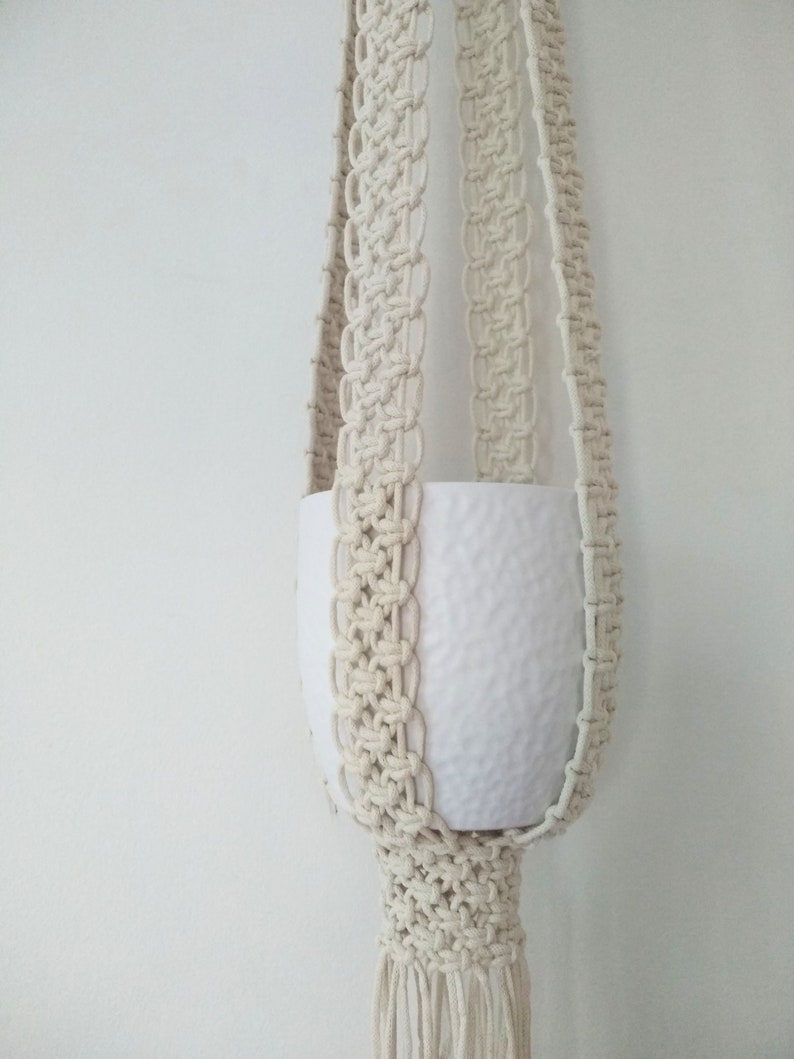 Pu&ograve; includere: Un portavasi in macram&egrave; bianco con un vaso in ceramica bianca. Il portavasi ha una frangia sul fondo.