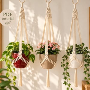 Puede incluir: Tres soportes de macramé para plantas en color blanco roto colgados de anillos de madera, con nudos intrincados y borlas. Sostienen una maceta roja con una planta colgante, una maceta beige con flores rosas y una maceta blanca con hiedra.