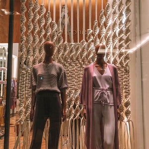 May include: Two mannequins are displayed in a storefront window. The mannequins are wearing different outfits. The mannequin on the left is wearing a gray sweater and black pants. The mannequin on the right is wearing a long, purple cardigan and light purple pants. The mannequins are standing in front of a wall made of macrame.