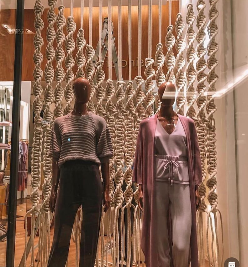 May include: Two mannequins are displayed in a storefront window. The mannequins are wearing different outfits. The mannequin on the left is wearing a gray sweater and black pants. The mannequin on the right is wearing a long, purple cardigan and light purple pants. The mannequins are standing in front of a wall made of macrame.