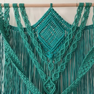 Op de afbeelding: Een turquoise macram&eacute; wandhanger met een ruitpatroon in het midden en franjes aan de zijkanten.