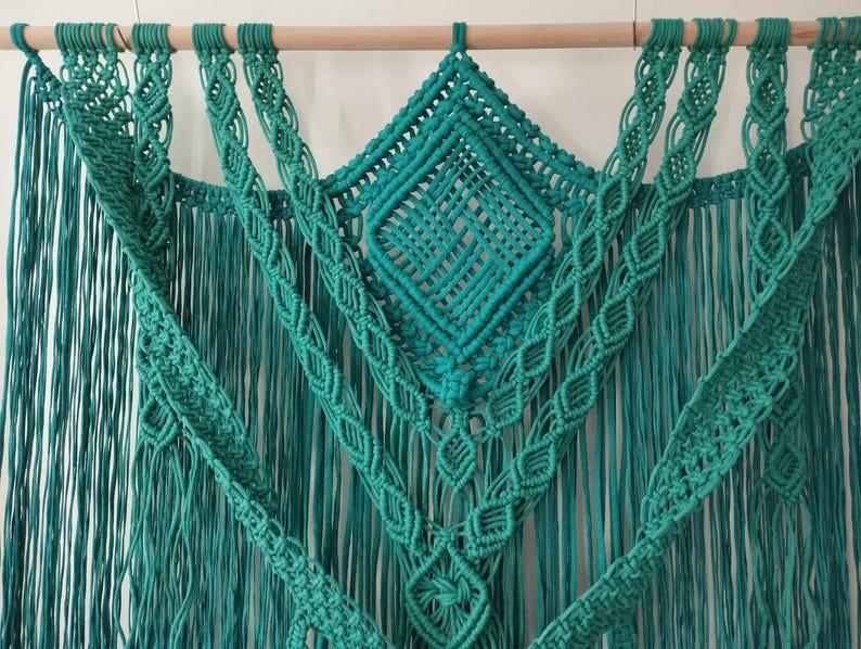 Op de afbeelding: Een turquoise macram&eacute; wandhanger met een ruitpatroon in het midden en franjes aan de zijkanten.