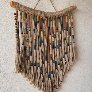 Pode incluir: Uma tapeçaria de parede feita de fibras naturais com um ramo de madeira no topo. A tapeçaria apresenta uma variedade de cordas coloridas com borlas na parte inferior.
